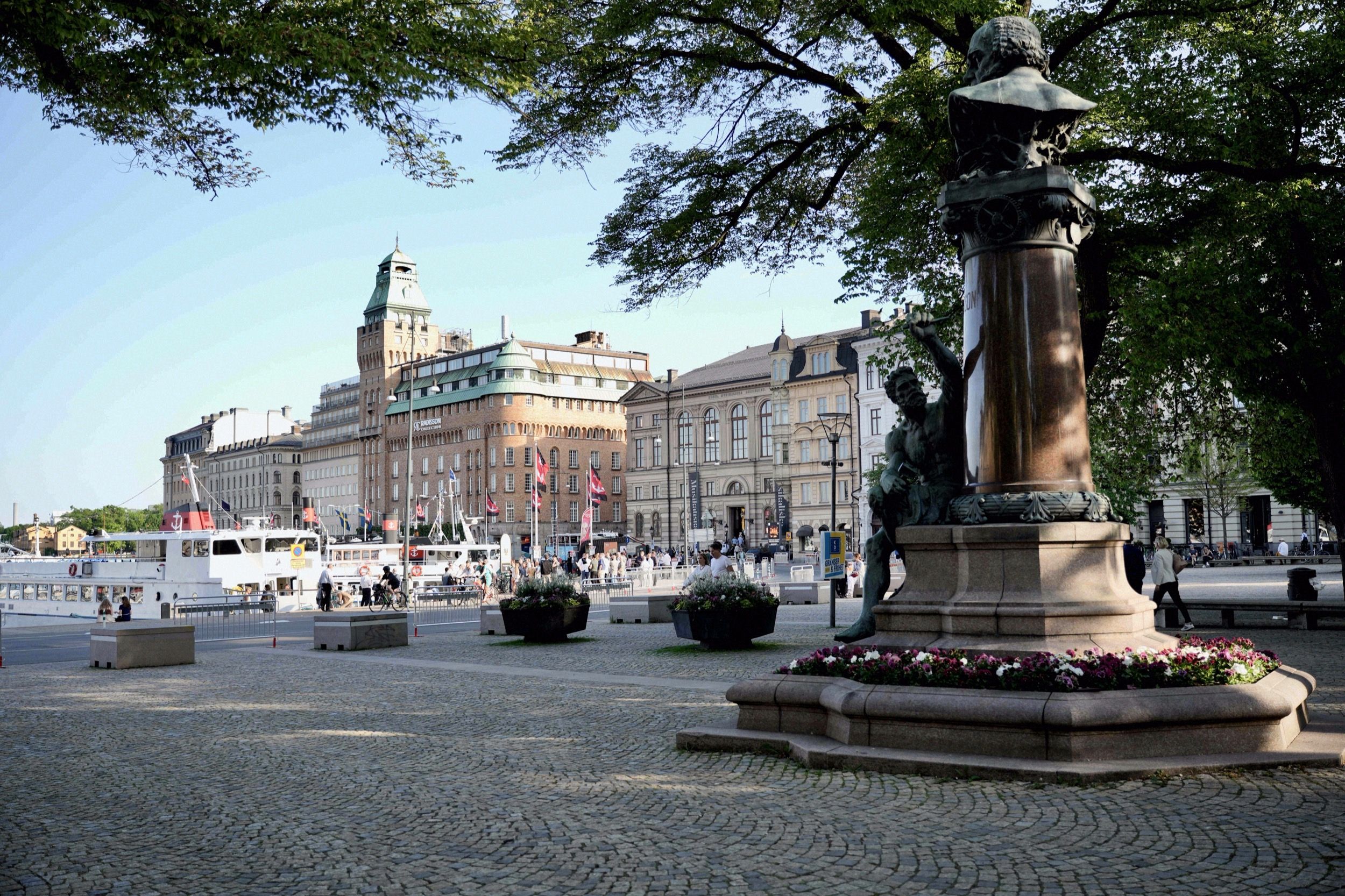 Blick über einen gepflasterten Platz mit Bronzebrunnen in Richtung Hafen und Hotel Grand Hôtel am Blasieholmen, im Abendsonnenlicht.