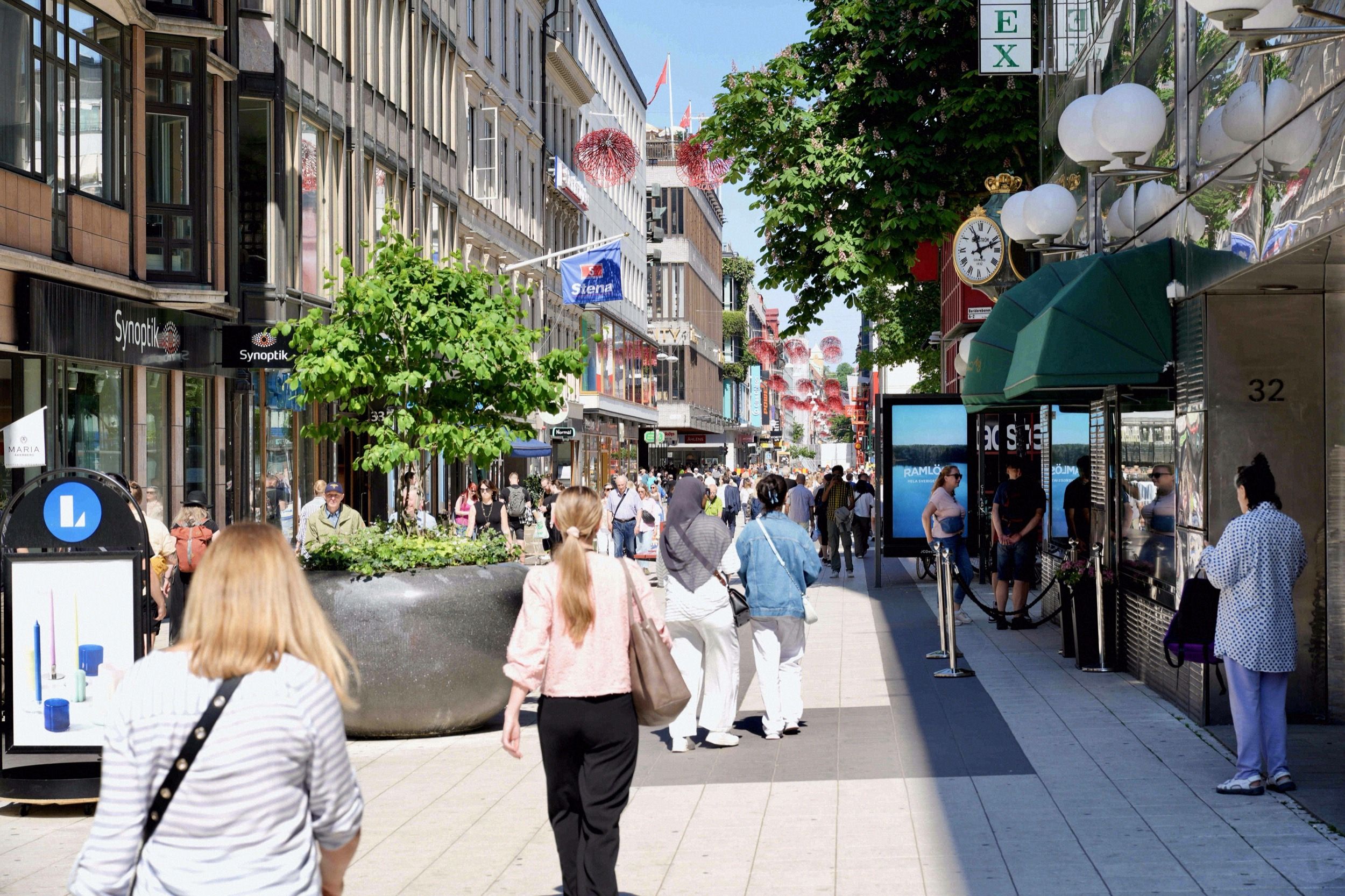 Belebte Drottninggatan (Fußgängerzone) mit Geschäften, Passanten und sommerlicher Dekoration.