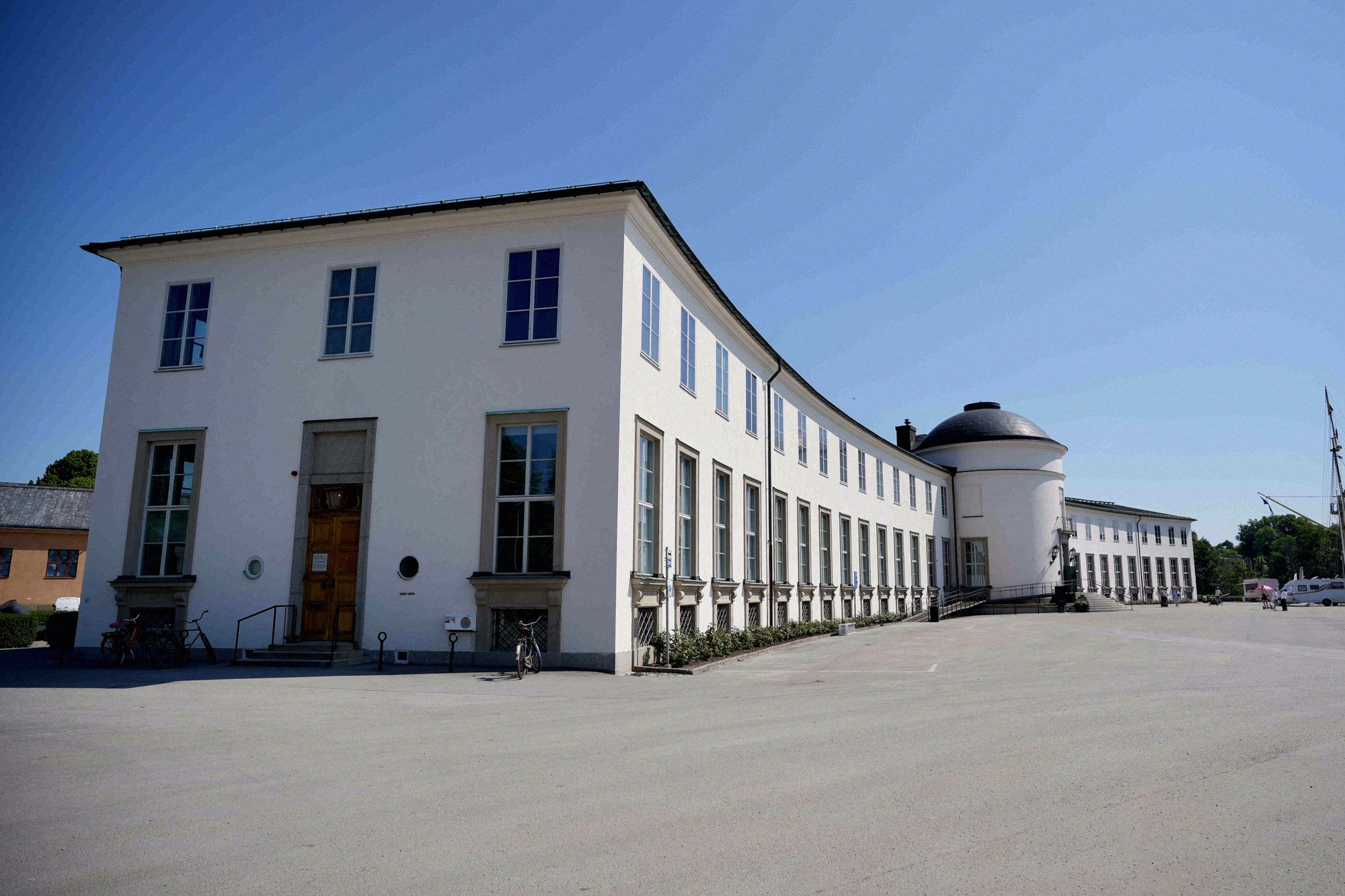 The neoclassical building of the Sjöhistoriska museet, with a historic sailing ship behind it. Side view of the long white neoclassical museum building with round tower and sailing ship